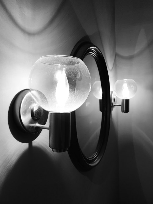 A modern wall sconce with a glass bulb, reflecting in a circular mirror, set against a textured wall.