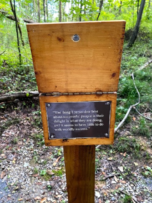 A wooden sign in a forest with a quote about success and delight in one's work.
