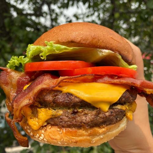 A hand holding a juicy burger with bacon, cheese, lettuce, and tomato, set against a blurred outdoor background.