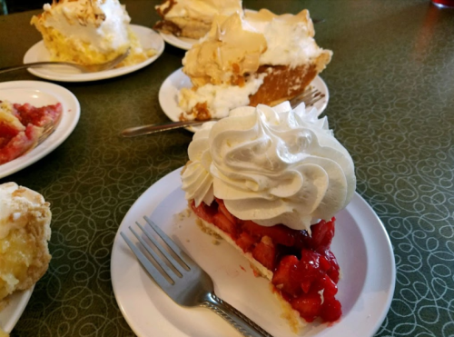 A slice of cherry pie topped with whipped cream, with other pie slices in the background on white plates.