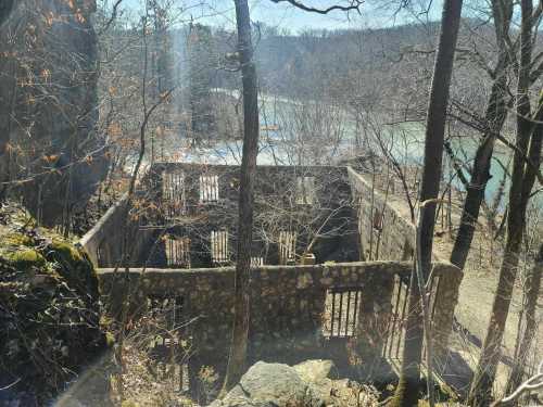 Ruins of a stone structure surrounded by bare trees, overlooking a river in a wooded area.