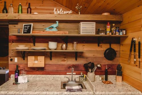 A cozy kitchen with wooden walls, shelves holding dishes, and a sink with utensils and bottles on the counter.