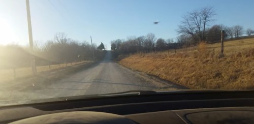 A view of a gravel road stretching into the distance, lined with trees and fields under a clear blue sky.