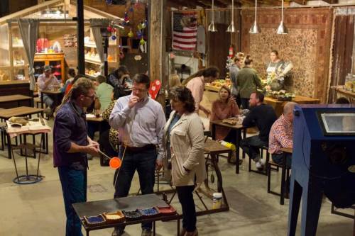 A lively indoor market scene with people socializing, dining, and browsing various stalls in a rustic setting.
