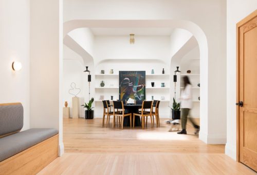 A bright, modern dining area with a large table, art on the wall, and a person walking by in a stylish home interior.