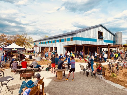 A lively outdoor gathering at a rustic building, with people socializing, biking, and enjoying the atmosphere.