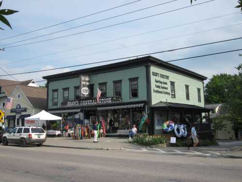 A green general store with a porch, flags, and colorful merchandise outside, set in a small town.