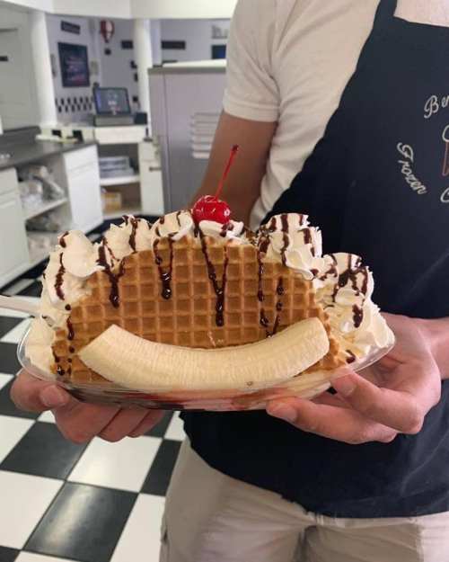 A person holds a dessert featuring a waffle topped with whipped cream, chocolate drizzle, a cherry, and a banana.
