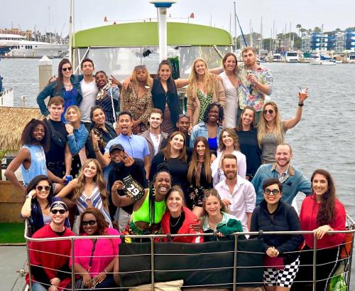 A large group of people posing together on a boat, with a waterfront and marina in the background.