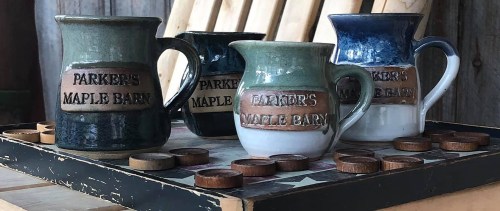 Four ceramic pitchers with "Parker's Maple Barn" labels, displayed on a wooden table with game pieces.