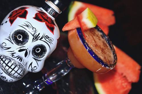 A decorative skull bottle beside a cocktail glass with a watermelon garnish and slices in the background.