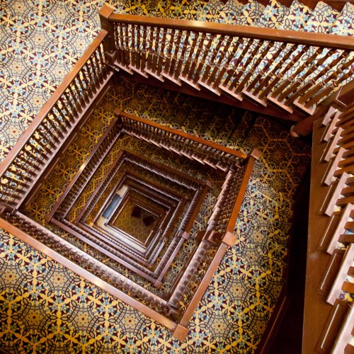 A spiral staircase with wooden railings, viewed from above, surrounded by patterned carpet.