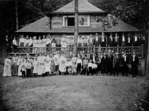 A large group of people, dressed in early 20th-century clothing, poses in front of a rustic wooden house.