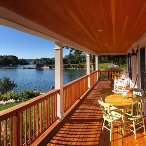 A scenic view of a lake from a porch with wooden flooring, chairs, and a table, surrounded by greenery and blue skies.