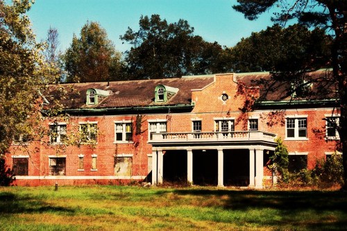 Abandoned brick building with a porch, surrounded by overgrown grass and trees under a clear blue sky.