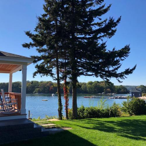 A serene lakeside view featuring a porch, rocking chairs, and lush greenery under a clear blue sky.