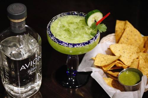 A green cocktail with cucumber and a red garnish, next to a bottle of tequila and a basket of tortilla chips with salsa.