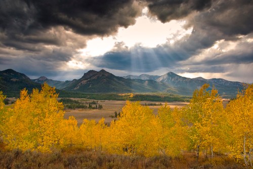 A dramatic landscape featuring golden autumn trees, mountains, and dark storm clouds with rays of sunlight breaking through.