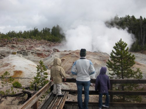 A person and two children stand on a wooden platform, gazing at a steaming geothermal area surrounded by trees.