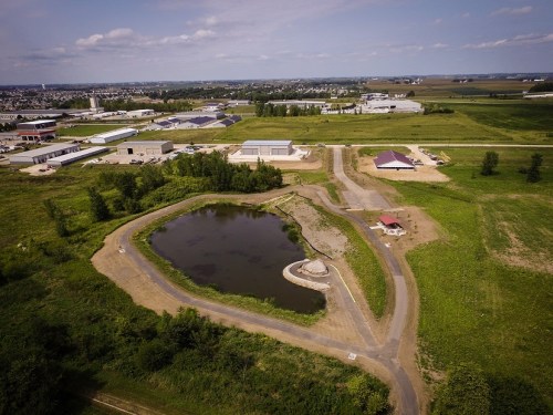 Aerial view of a pond surrounded by greenery and industrial buildings in the background under a clear sky.