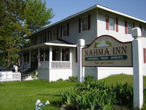 A white inn with red shutters and a sign reading "Nahma Inn" surrounded by greenery and a picket fence.