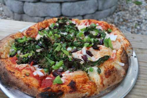 A freshly baked pizza topped with green peppers, onions, olives, and leafy greens on a wooden table.