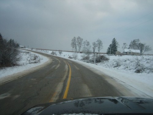 A winding road covered in snow, flanked by bare trees and a distant house under a cloudy sky.