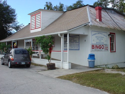 A small, white building with a sloped roof, featuring signs for bingo and educational activities, parked car nearby.