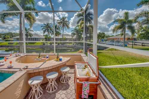 A sunny outdoor patio with a pool, bar area, and palm trees overlooking a canal. Bright blue sky and fluffy clouds.