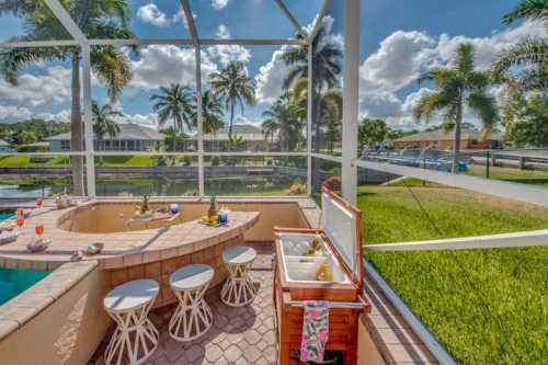 A sunny outdoor patio with a pool, bar area, and palm trees overlooking a canal. Bright blue sky and fluffy clouds.