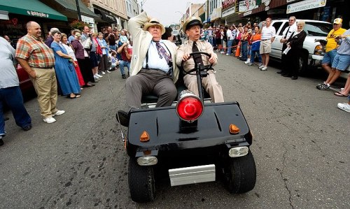 Two men in a small vehicle, one wearing a suit and the other in a police uniform, driving down a crowded street.