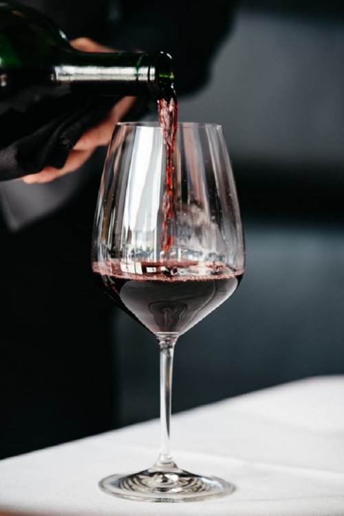 A hand pours red wine into a large glass, set on a white tablecloth.
