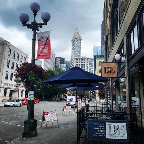 Street view of Pioneer Square in Seattle, featuring a restaurant patio, lampposts, and city skyline under cloudy skies.