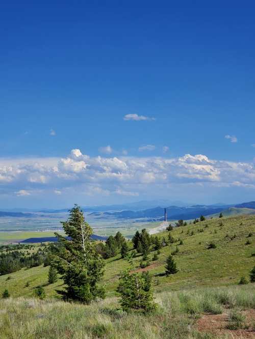 A scenic landscape featuring rolling green hills, scattered trees, and a blue sky with clouds in the distance.
