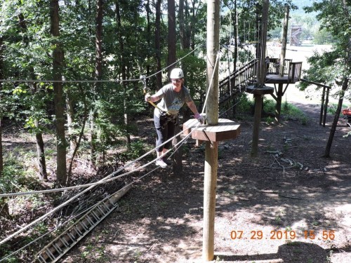 A person navigates a ropes course in a wooded area, balancing on a cable with a harness and safety gear.