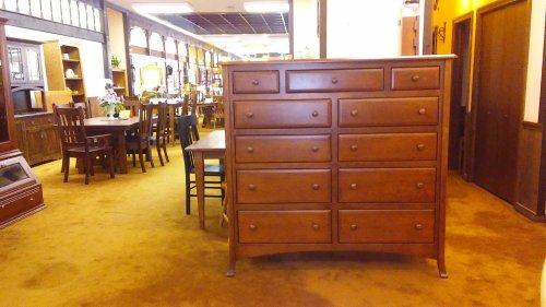 A wooden dresser with multiple drawers stands in a furniture showroom with tables and chairs in the background.