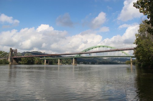 A green arch bridge spans a river, with a suspension bridge nearby, under a partly cloudy sky.