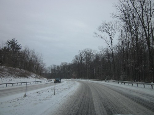 A snowy road curves through a forest, with a car driving on the right side and trees lining the roadside.