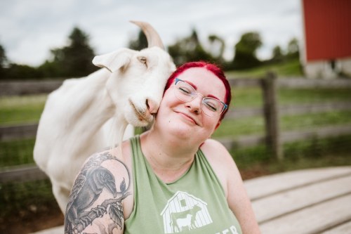 A smiling person with red hair and glasses poses with a goat nuzzling their shoulder in a farm setting.