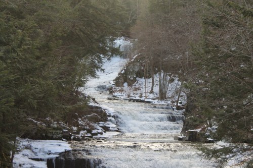 A serene waterfall cascades through a forest, surrounded by trees and snow, reflecting sunlight on the water's surface.