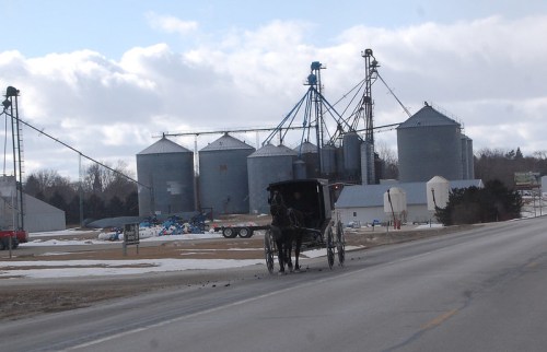 A horse-drawn buggy travels along a road near large grain silos under a cloudy sky.
