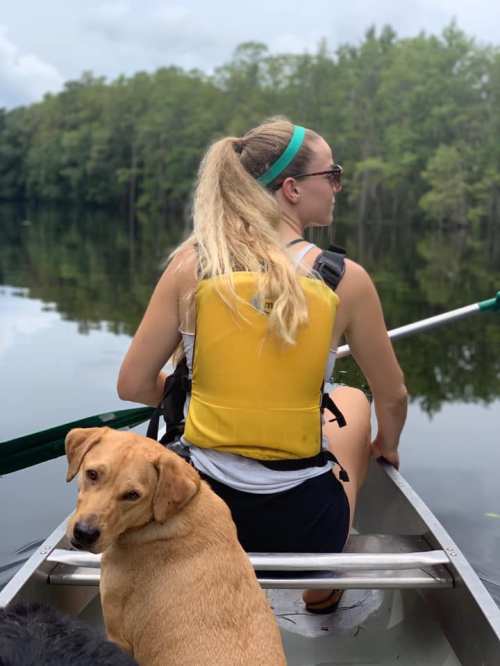 A person in a yellow life jacket paddles a canoe with a dog resting beside them on a calm lake surrounded by trees.