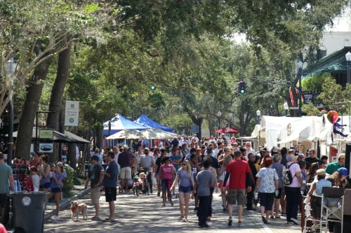 A busy street filled with people, tents, and trees, creating a lively outdoor market atmosphere.