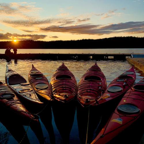 A serene sunset over a dock with kayaks lined up in the foreground and two people silhouetted in the background.