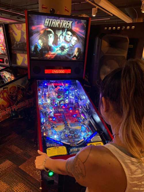 A person plays a Star Trek pinball machine, illuminated with colorful lights in an arcade setting.
