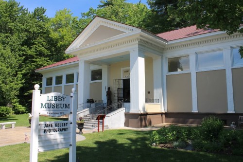 Exterior view of the Libby Museum, featuring a sign for Jane Kelley Photography and surrounded by greenery.