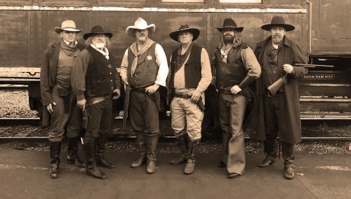 A group of six men dressed in Western attire, posing in front of a vintage train.