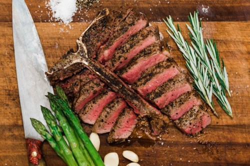 Sliced steak on a wooden board, garnished with rosemary, asparagus, and garlic, alongside a large knife.