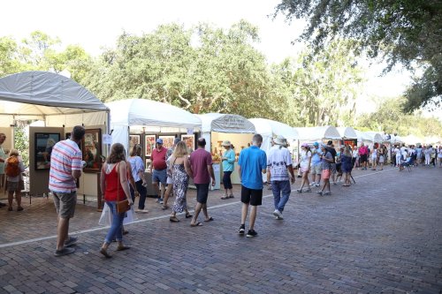 A bustling outdoor art fair with people browsing booths under white tents along a tree-lined path.