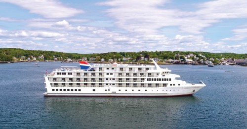 A large white cruise ship sails in calm waters, surrounded by green hills and a small coastal town in the background.